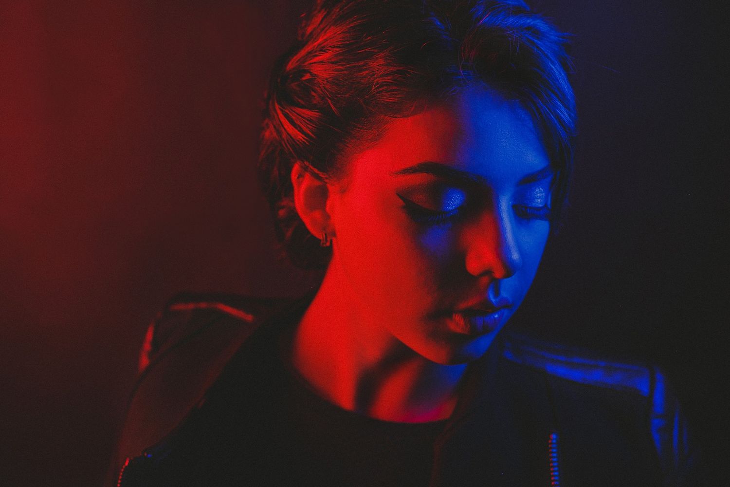 A young woman bathed in dramatic red and blue neon lights poses with closed eyes against a dark background.