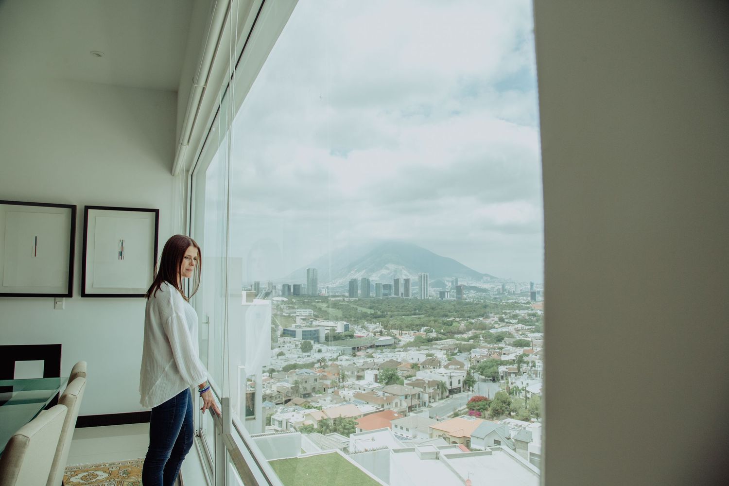 Woman in white blouse stands by floor-to-ceiling windows overlooking a city skyline with mountains in the background.