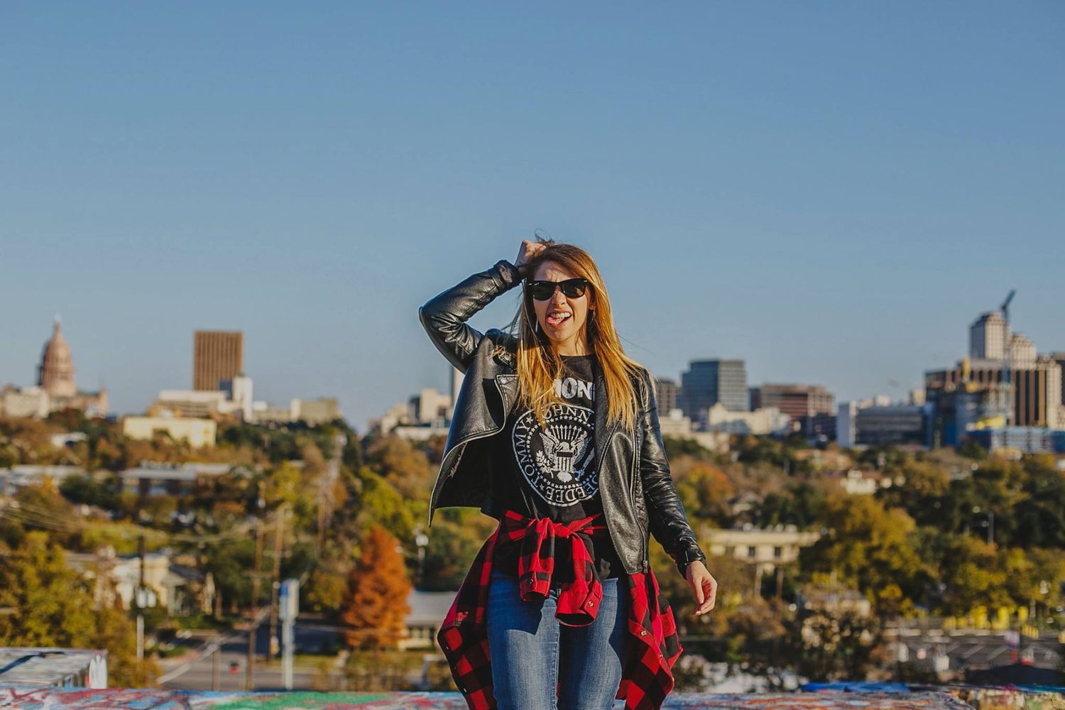 Woman in leather jacket and sunglasses smiling on rooftop with city skyline and blue sky in background.