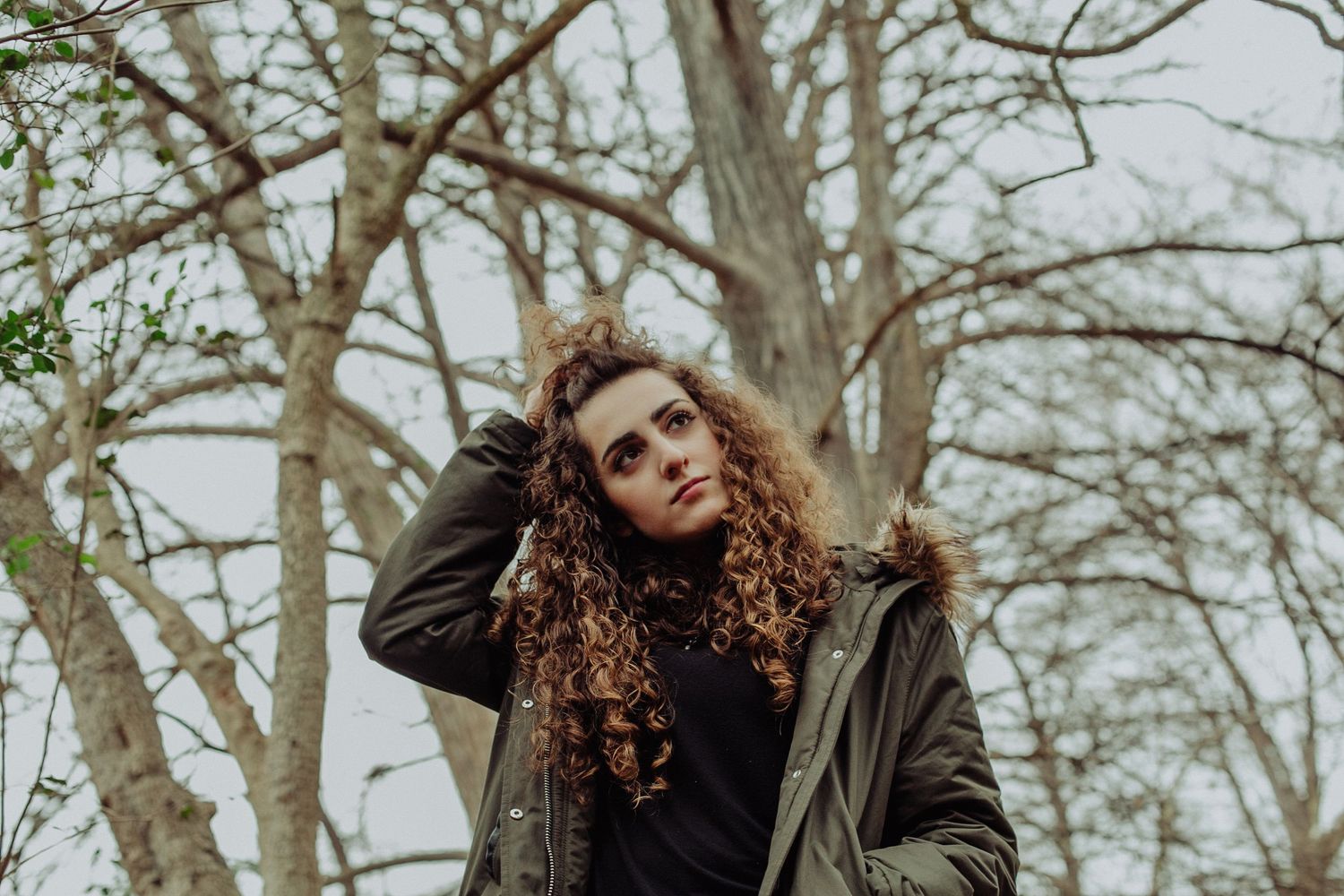 Young woman with curly hair poses outdoors in a green parka jacket among bare winter trees.