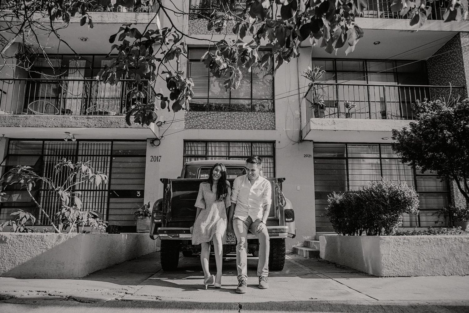 Black and white photo of a couple leaning against a vintage truck in front of a multi-story building on a city street.