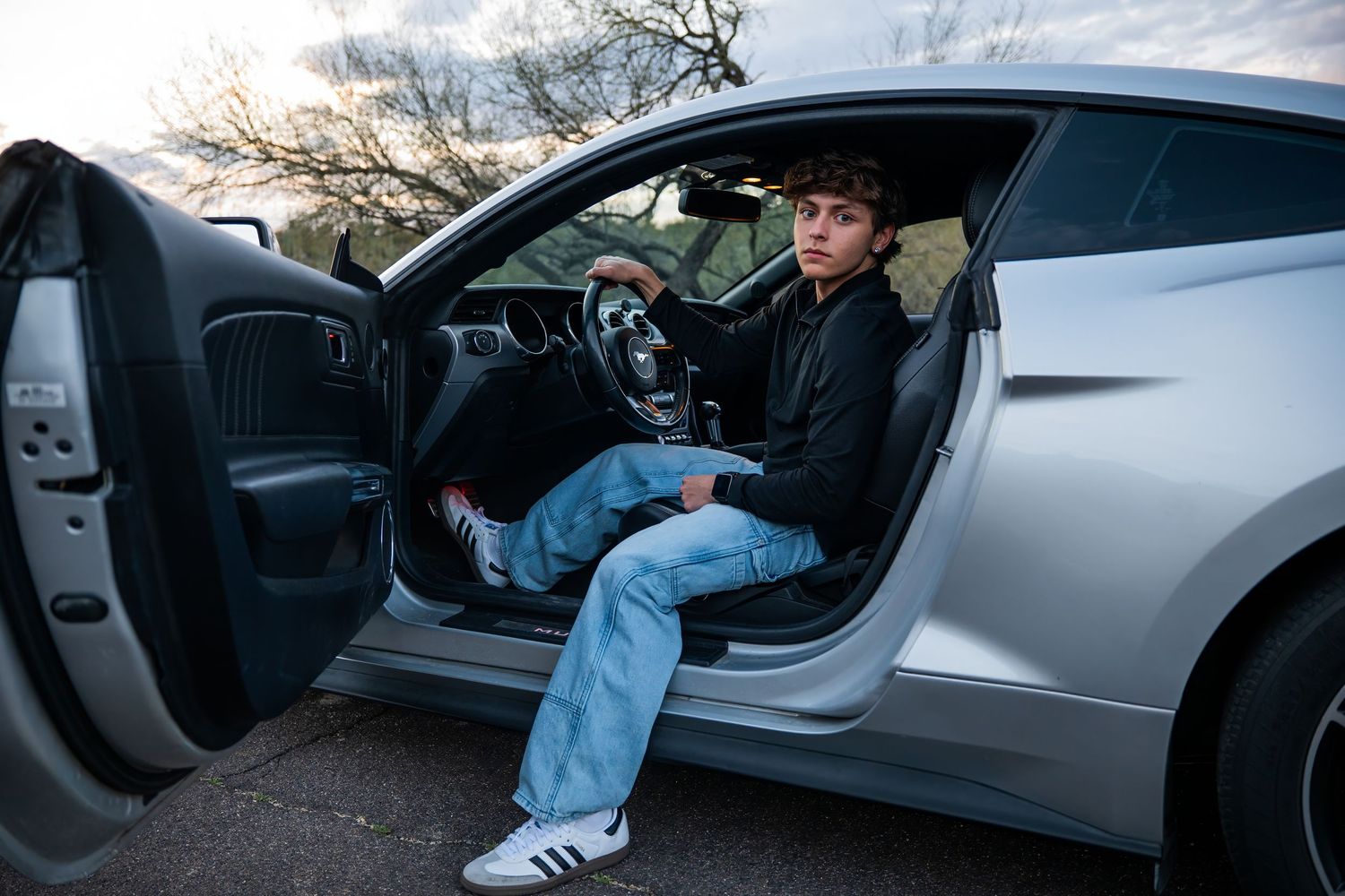Young man in jeans and white sneakers sitting in open door of silver sports car outdoors.