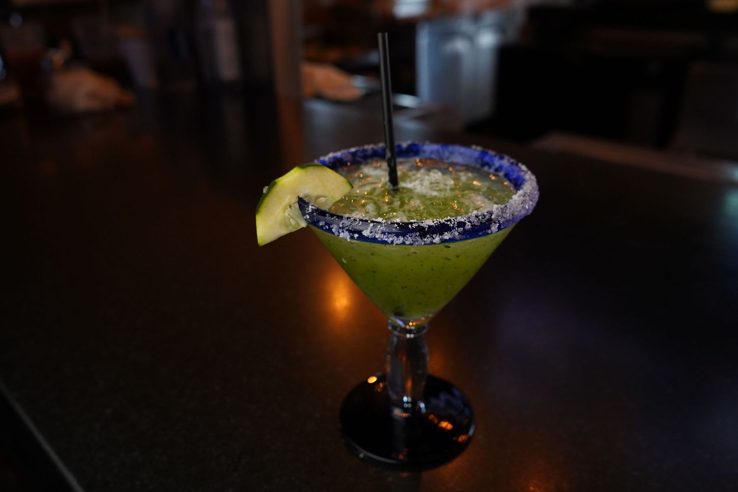 Green margarita in a salted rim glass with lime wedge and black straw on a dark bar.