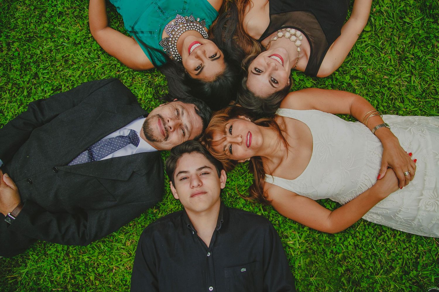 Five people lying on green grass in a circle with heads together, smiling upward, dressed in formal attire.