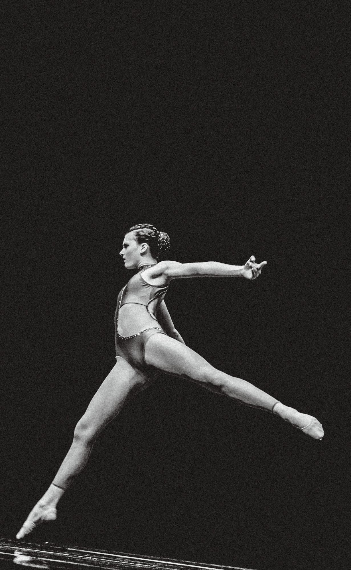 Black and white photo of a female ballet dancer mid-leap in a leotard, arms extended gracefully on a dark stage.