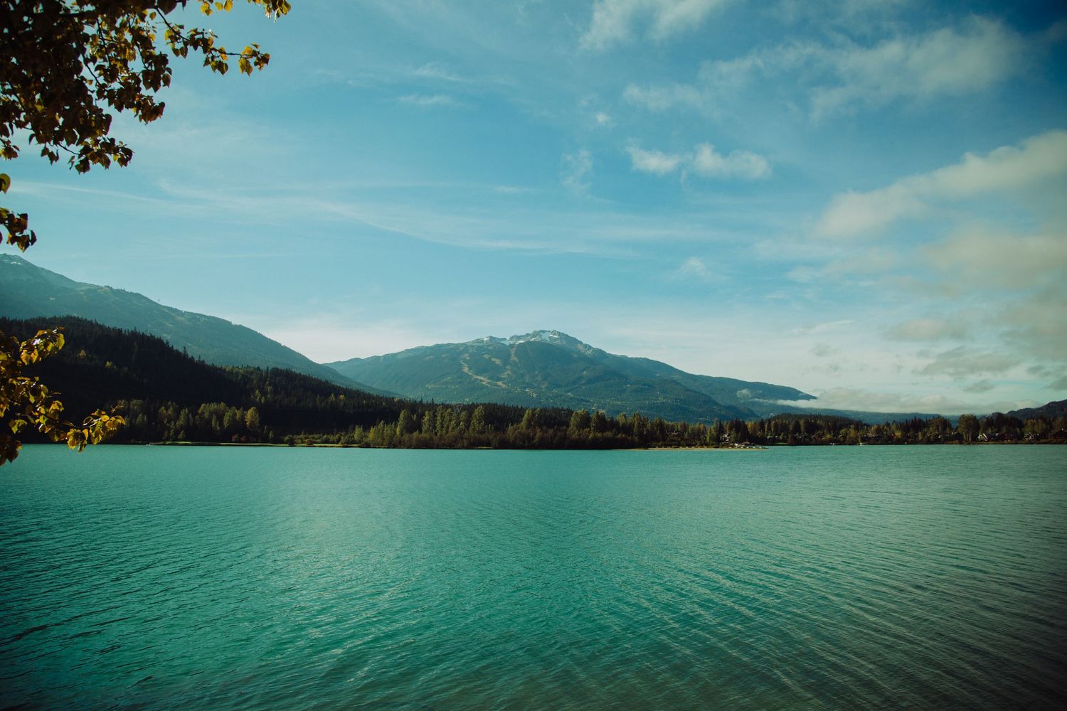 Serene turquoise mountain lake surrounded by autumn forest trees with snow-capped peaks and blue sky in background.