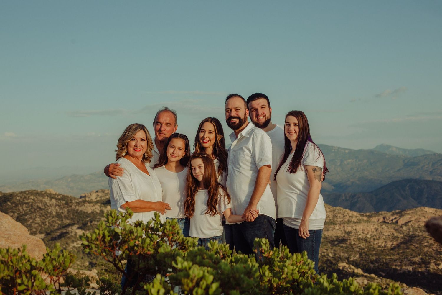 Family of eight posing together in white outfits on a scenic mountain overlook at sunset.