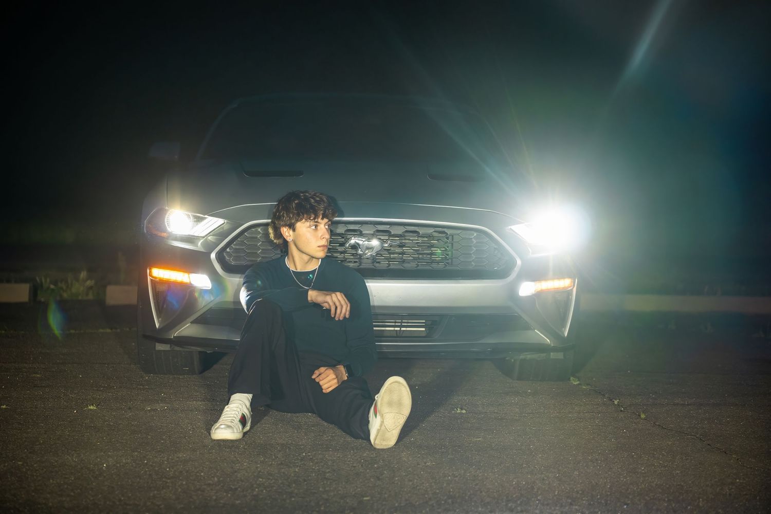 Young man sitting on pavement in front of illuminated car headlights at night wearing dark outfit and white sneakers.