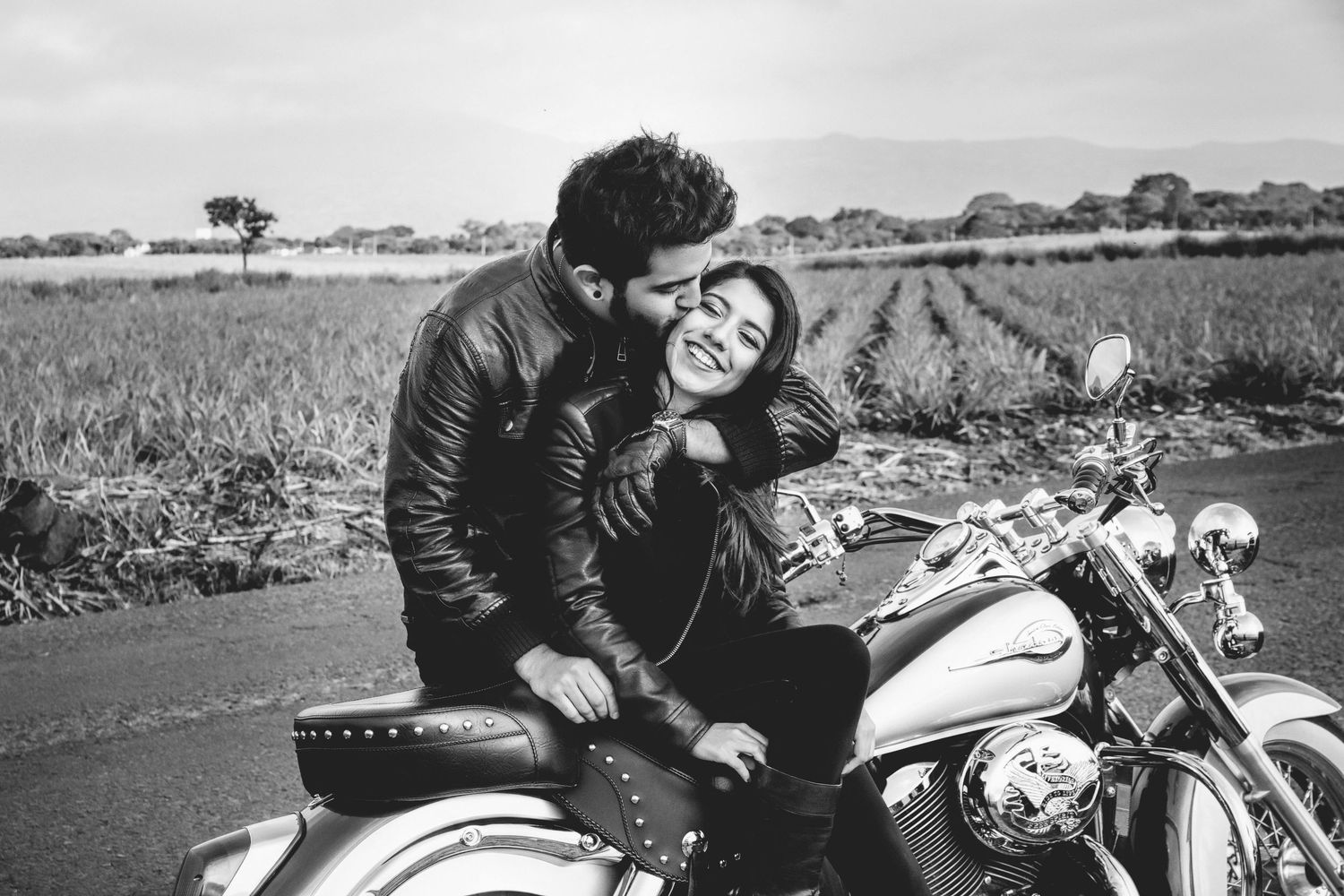 Black and white photo of a romantic couple in leather jackets sharing a tender moment on a classic motorcycle by a field.