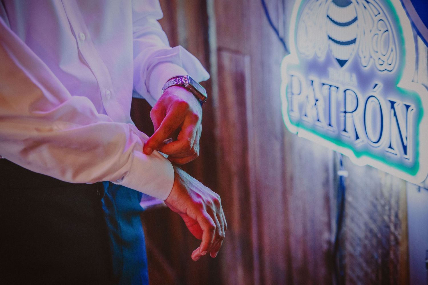 Man in white dress shirt adjusting cufflinks near a Patron tequila sign at an event.