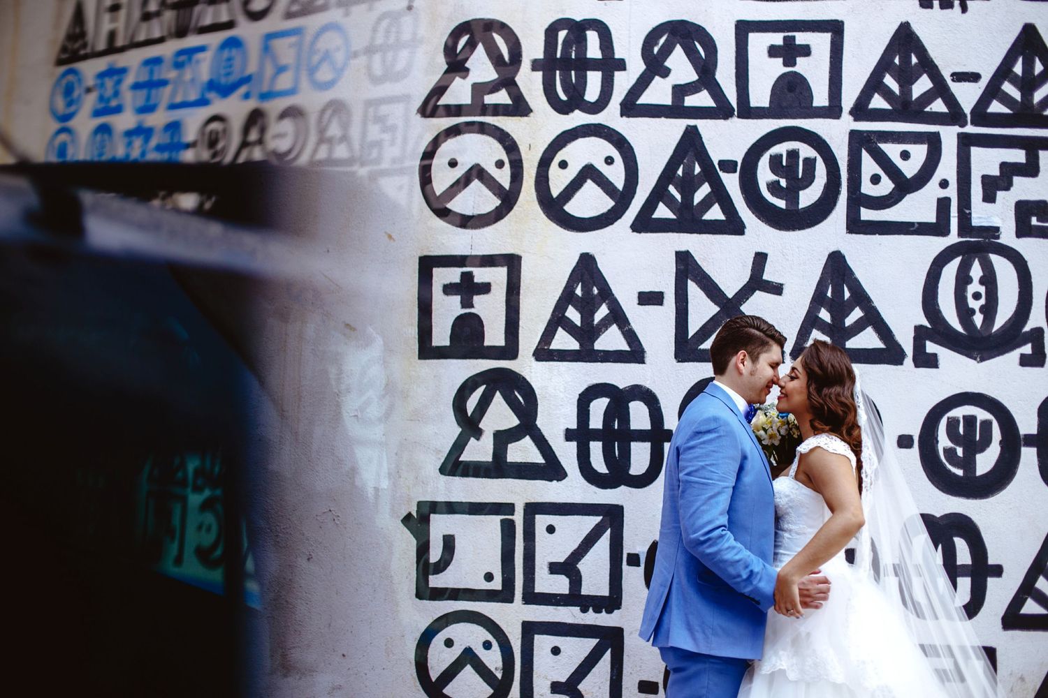 Newlyweds share a romantic moment in front of a bold geometric graffiti mural, bride in white gown and groom in blue suit.