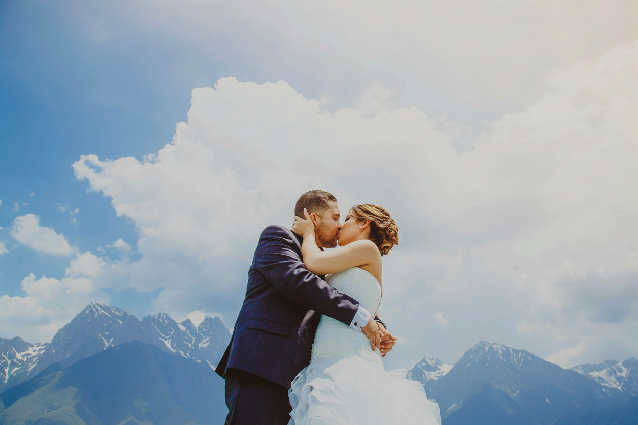 A couple shares a romantic kiss in wedding attire with stunning mountain peaks and clouds in the background.