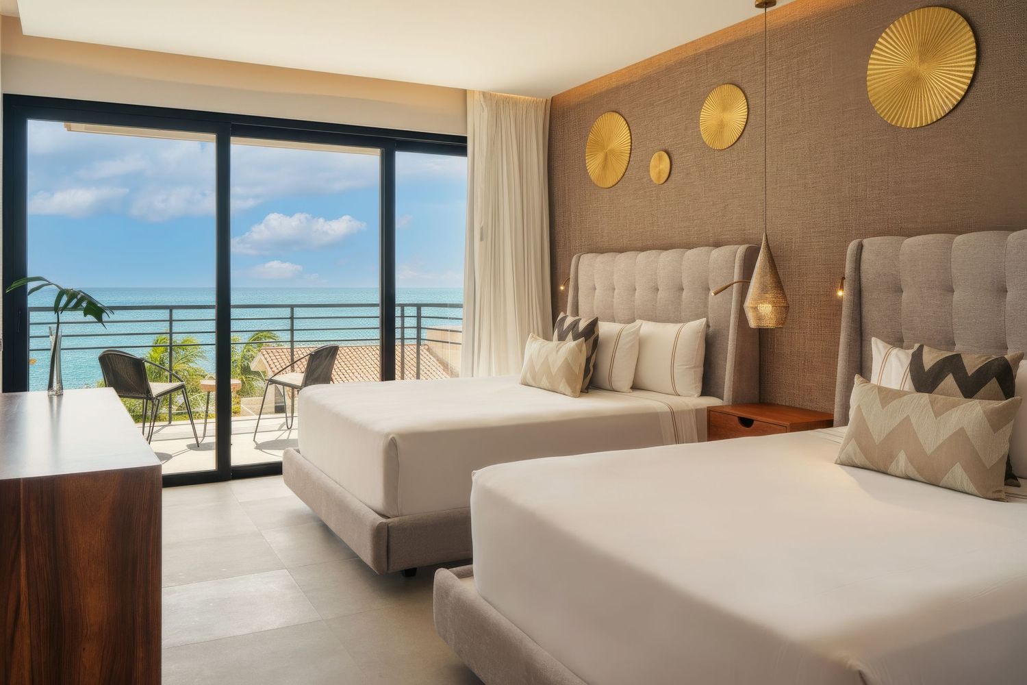 Elegant hotel room with two queen beds, neutral decor, gold wall accents, and a balcony overlooking a turquoise ocean view.