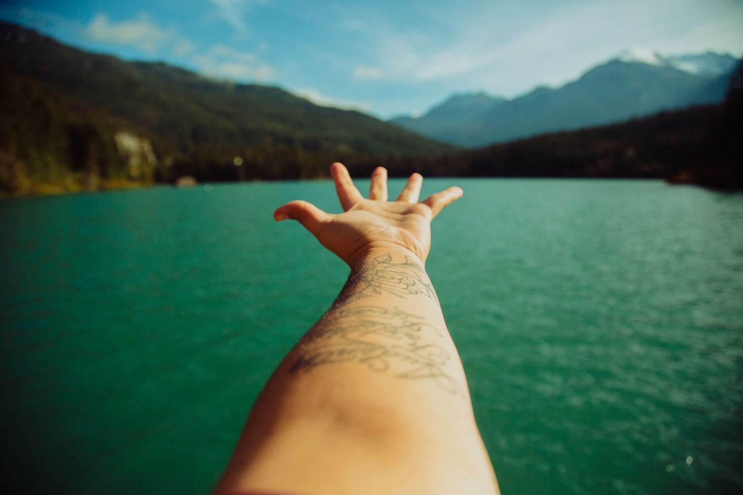 Tattooed arm reaching toward turquoise lake with mountains in background.