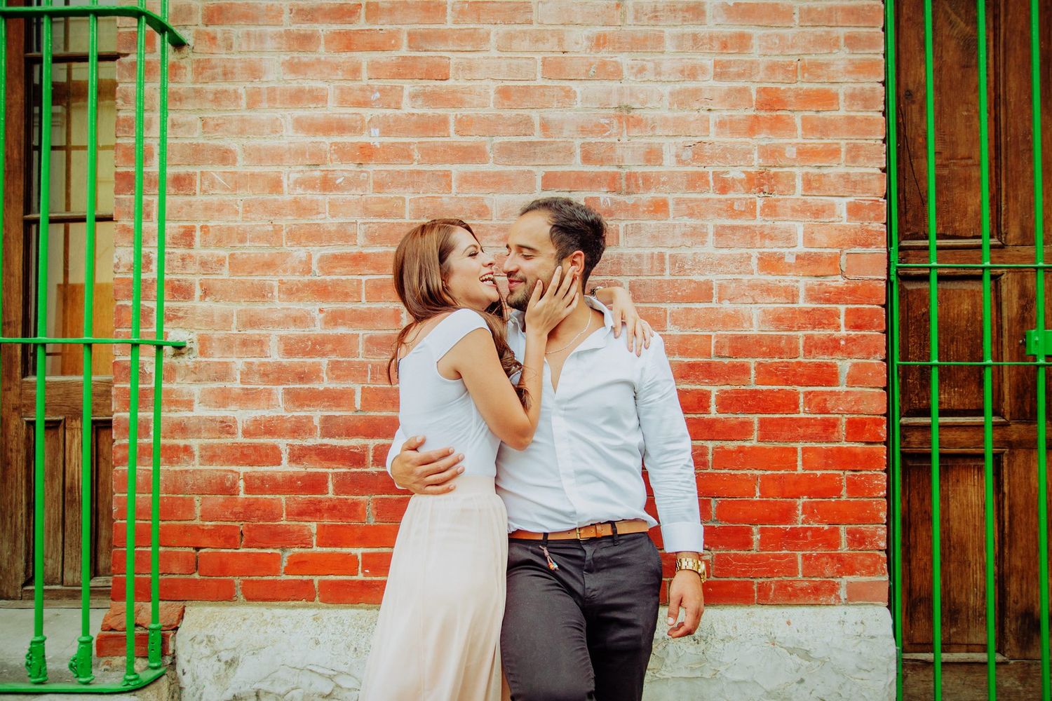 A couple shares a romantic kiss against a rustic red brick wall with green metal doors.