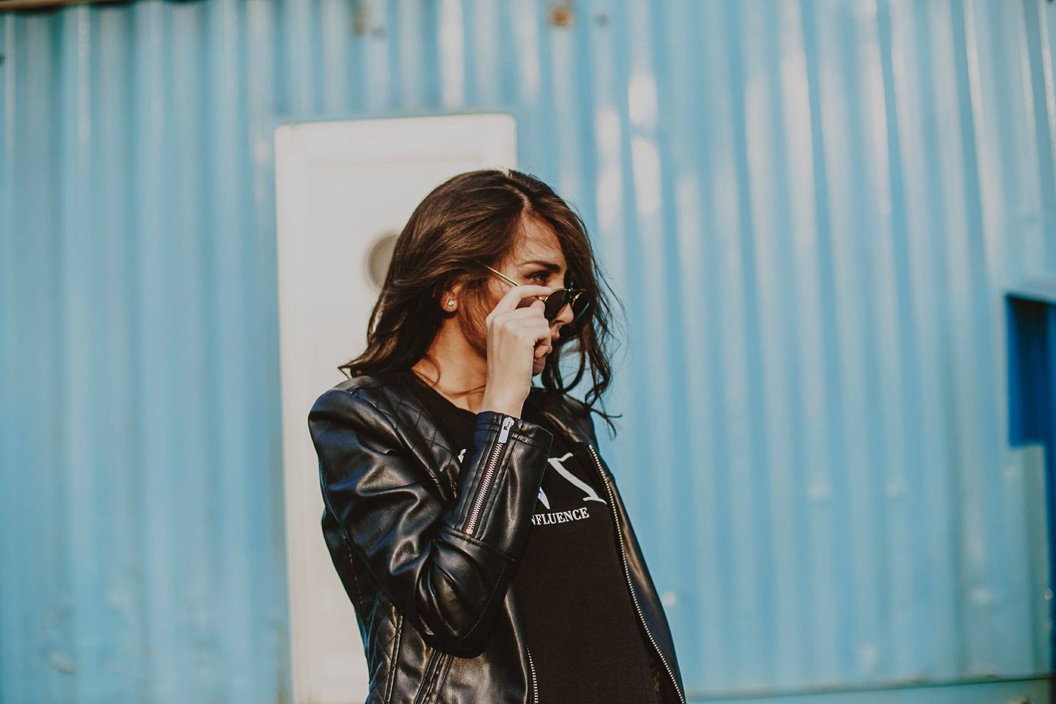 A young woman in a black leather jacket adjusts her sunglasses outdoors near a blue corrugated metal wall.