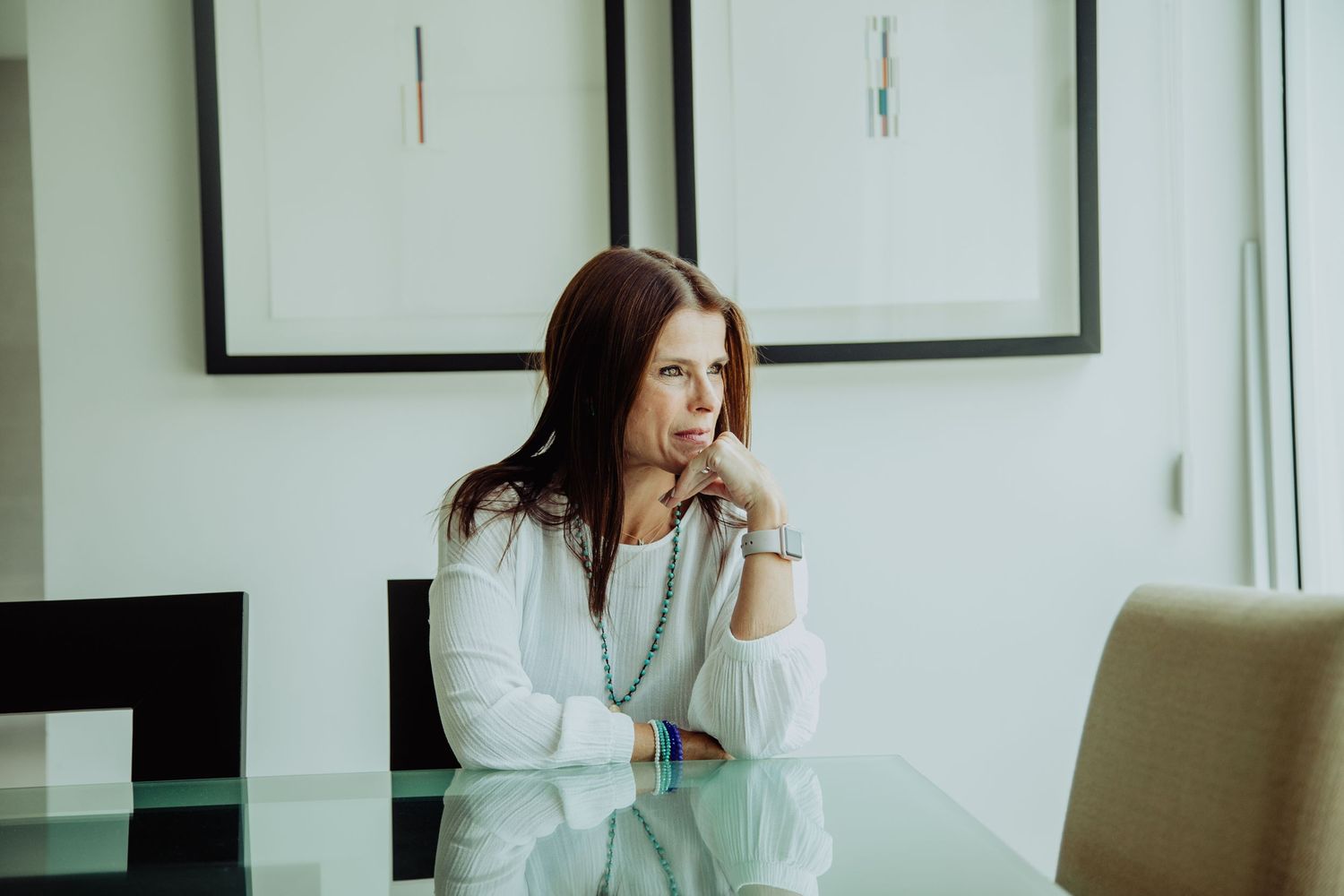 Woman in white blouse sitting at glass table, resting chin on hand, looking thoughtfully to the side in a modern room.