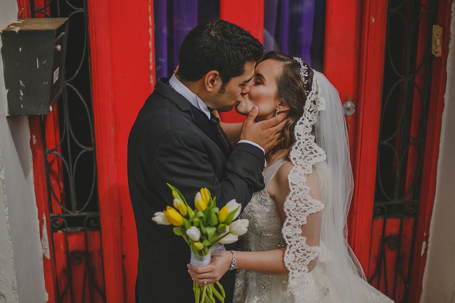 Groom tenderly holds bride's face near a vibrant red door as she holds yellow tulip bouquet.