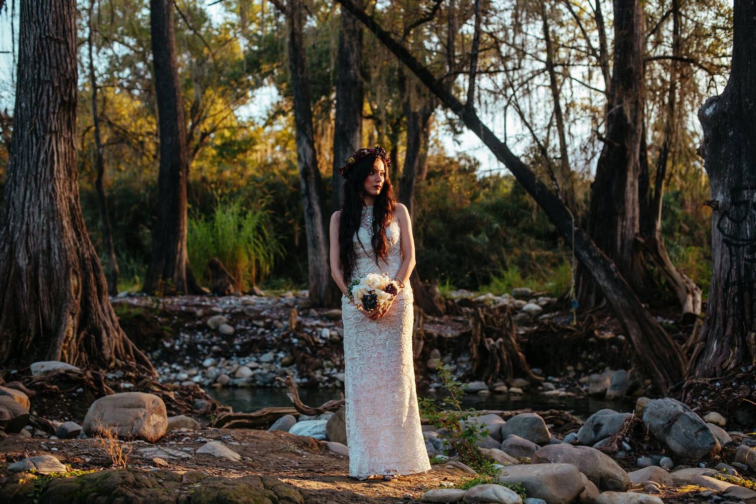 Bride in a white lace gown holds a bouquet while standing among trees and rocks at sunset.