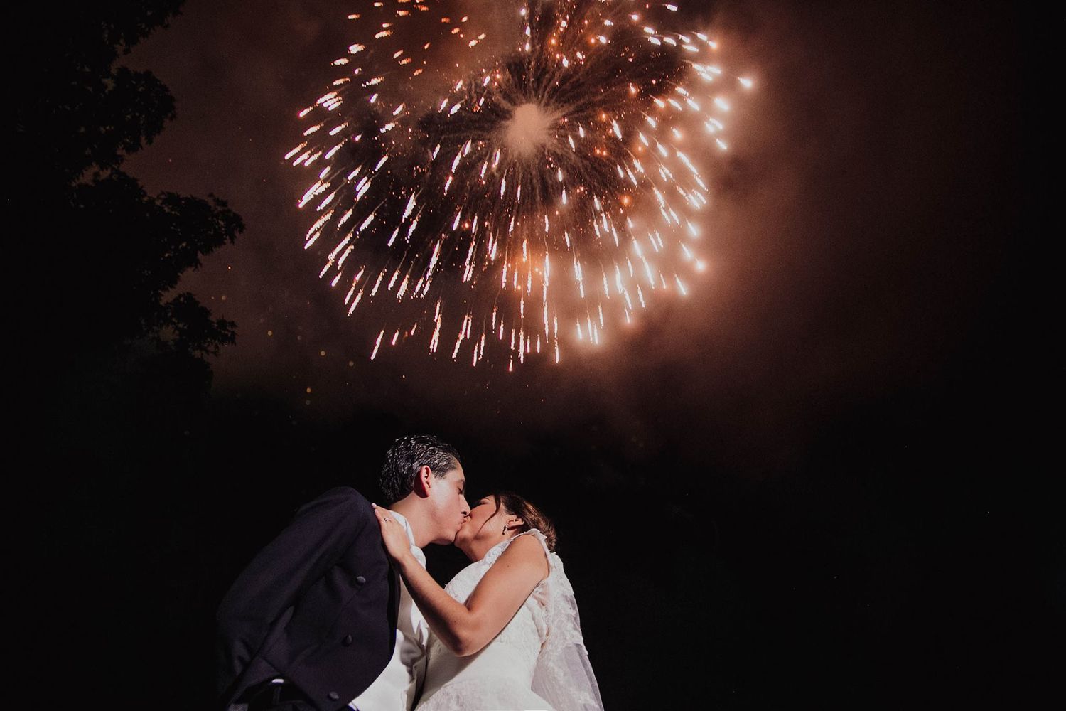 Bride and groom share a romantic kiss under a stunning fireworks display lighting up the dark night sky.