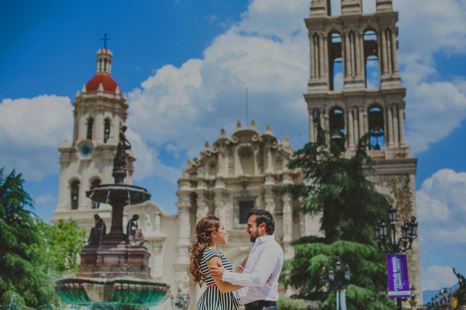 Couple embraces near ornate fountain with historic Mexican cathedral and bell towers under blue sky.