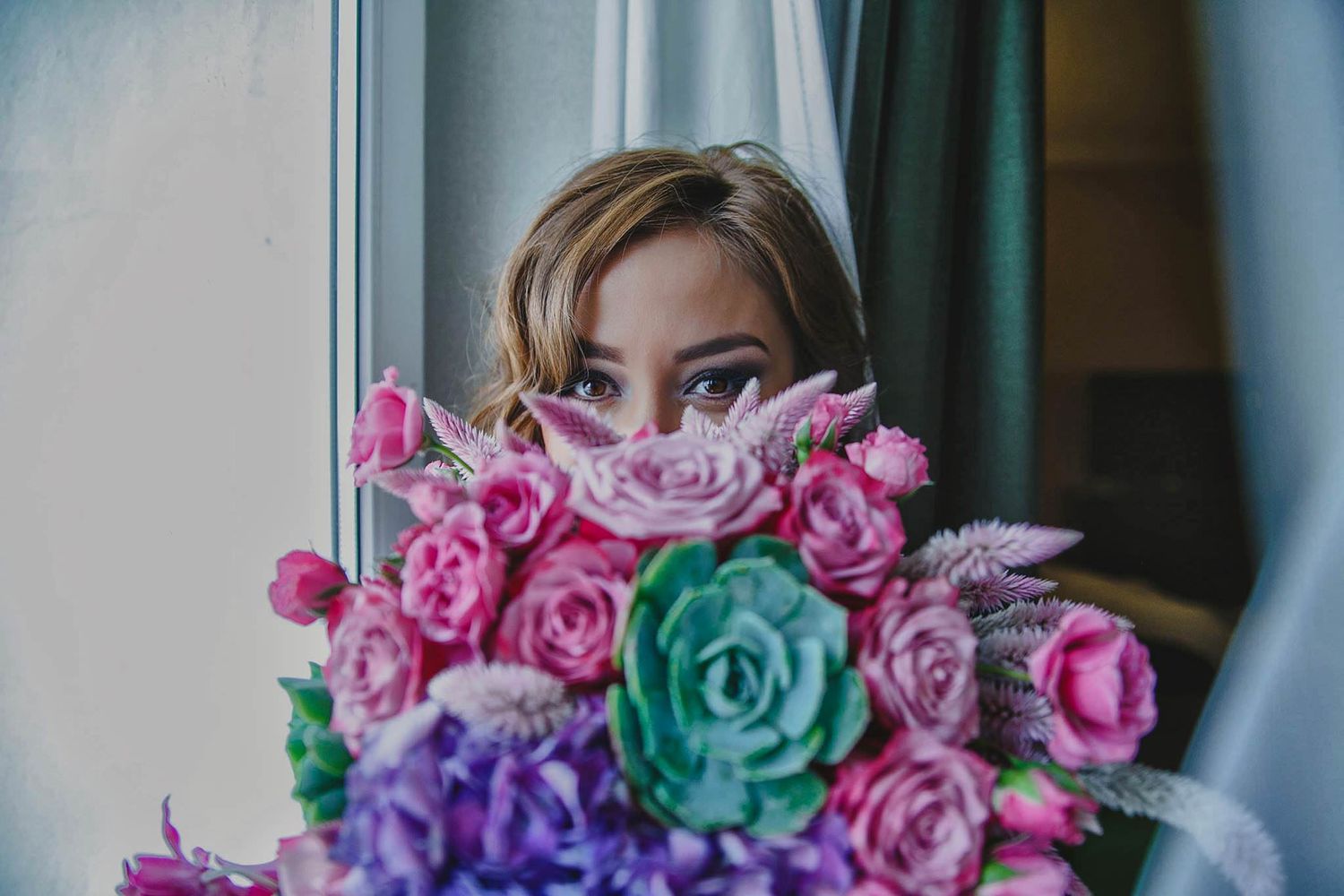 A bride peeks over a vibrant bouquet of pink and teal flowers near a window.
