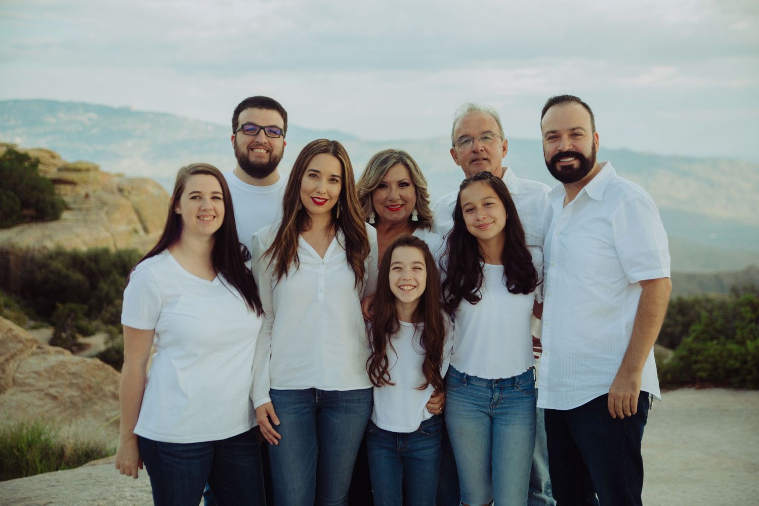 Family of eight smiling together outdoors in white shirts and jeans with a scenic mountain landscape in the background.