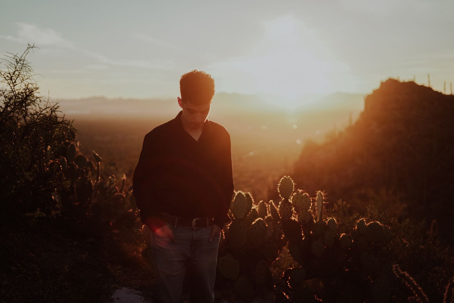 Man standing among cacti at golden hour sunset with mountains and glowing orange sky in the background.