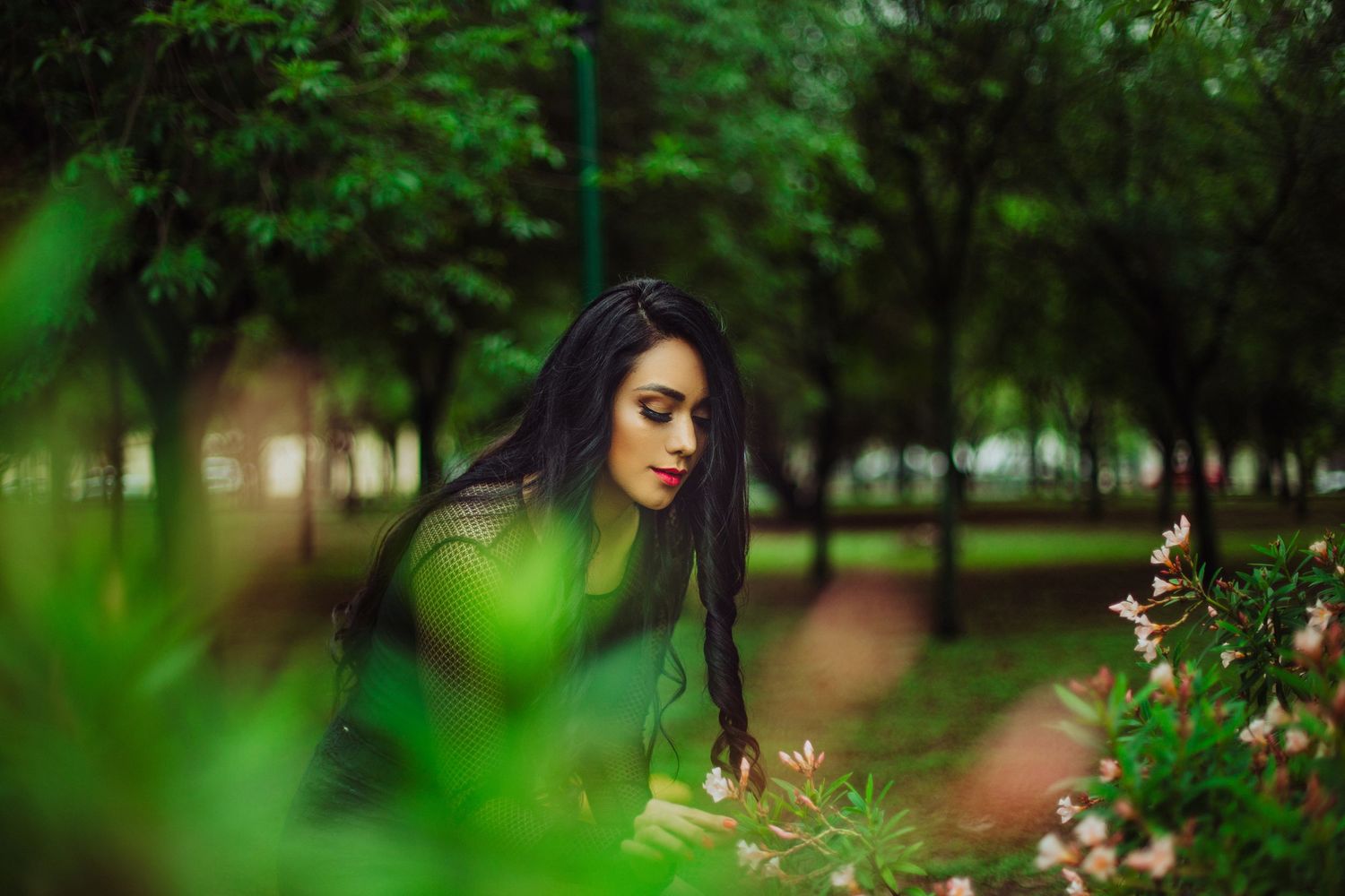 A young woman with long dark hair and red lips gazes down at blooming flowers in a lush green park.