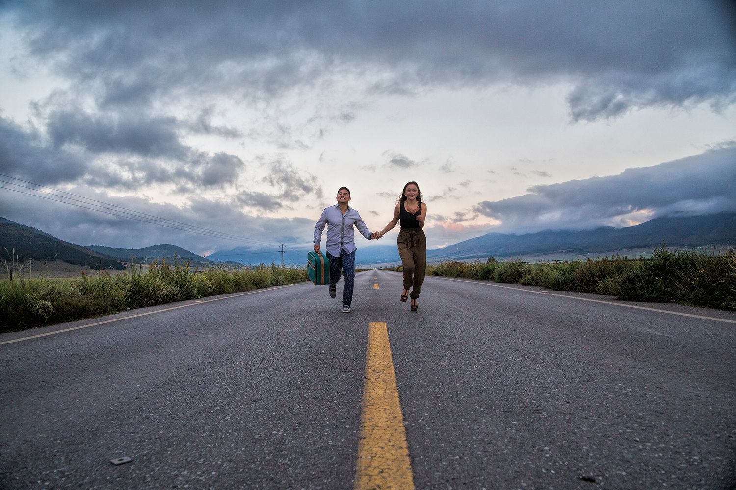 Couple holding hands running down an empty road with mountains and dramatic cloudy sky at sunset.