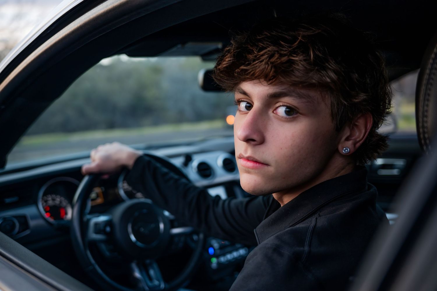 Young man with curly hair sitting in a car, gripping the steering wheel, wearing a black jacket, looking at the camera.