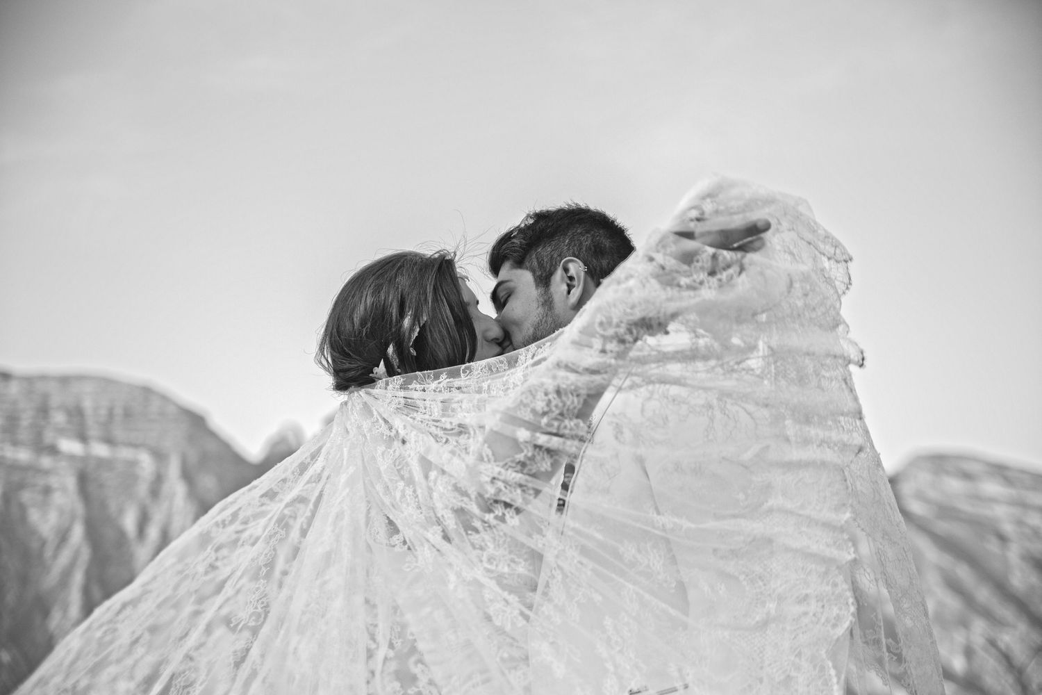 Black and white photo of a couple sharing a kiss wrapped in a flowing lace wedding veil outdoors.