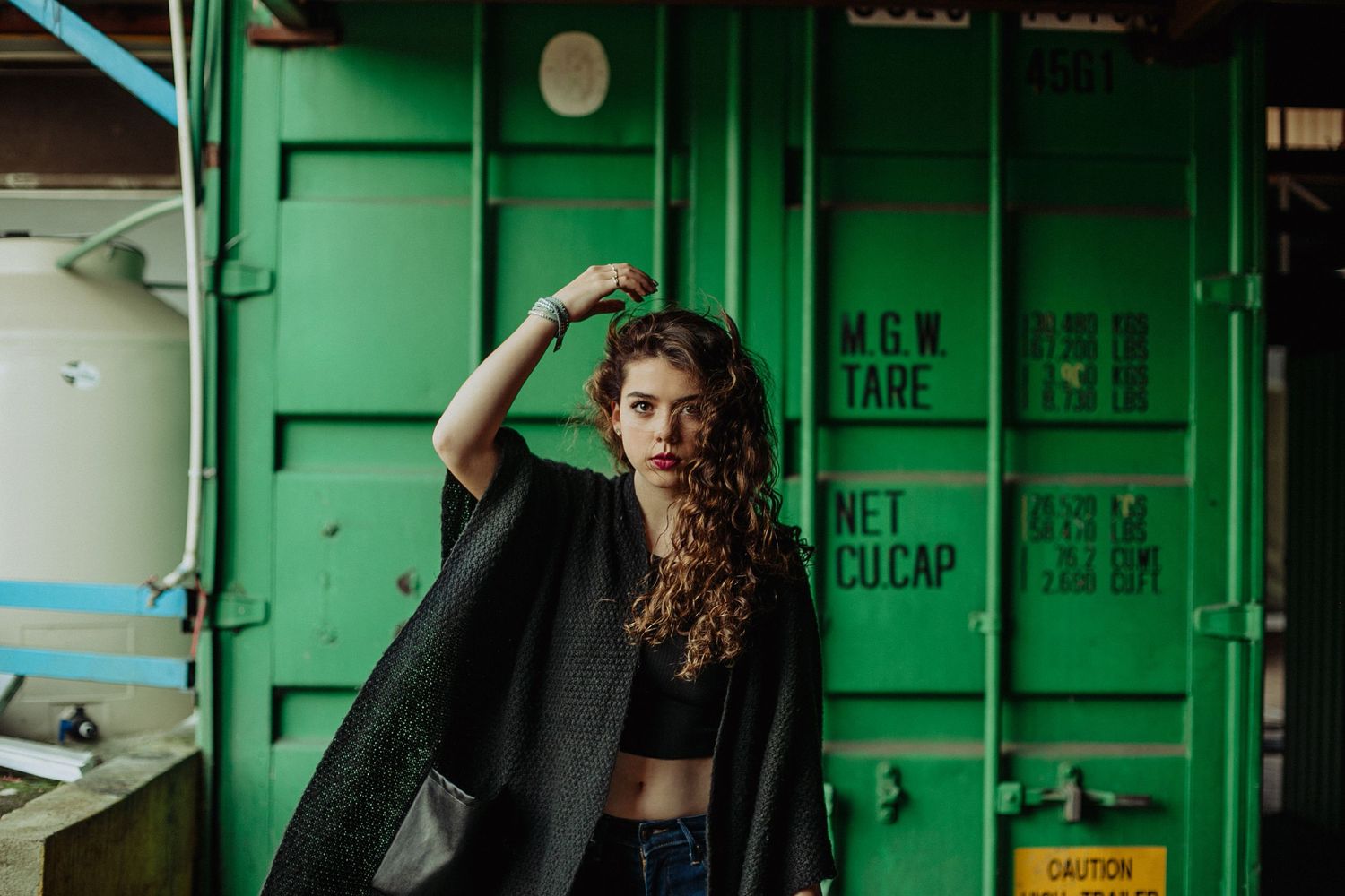 Young woman with curly hair wearing a black cape poses in front of a green shipping container.
