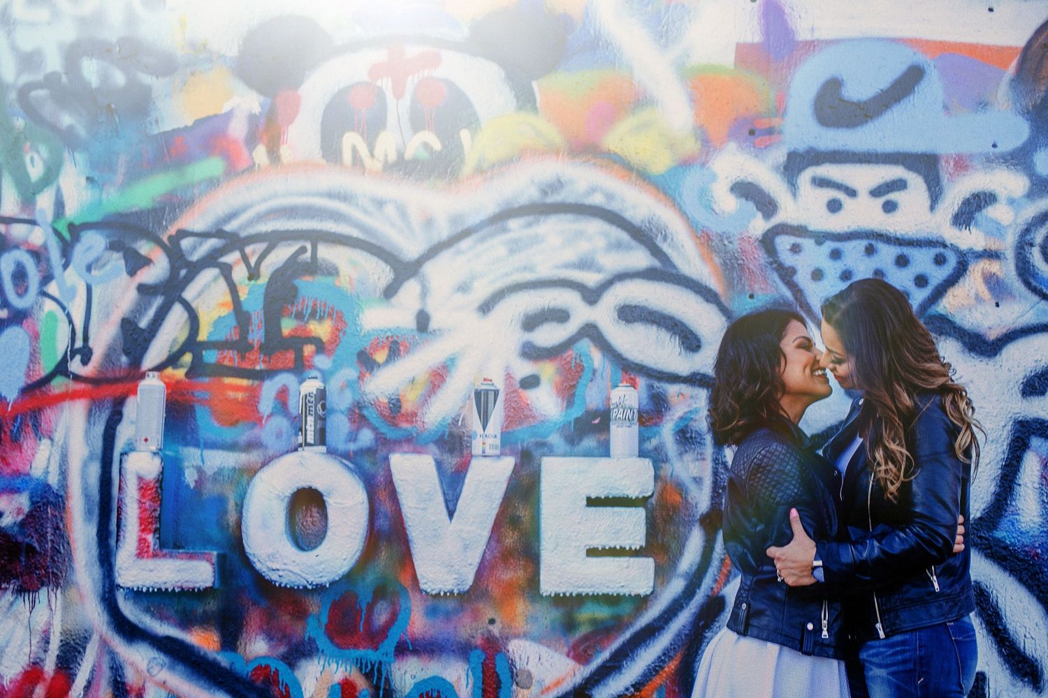 Two women sharing a kiss in front of a colorful graffiti wall featuring the word LOVE in white letters.