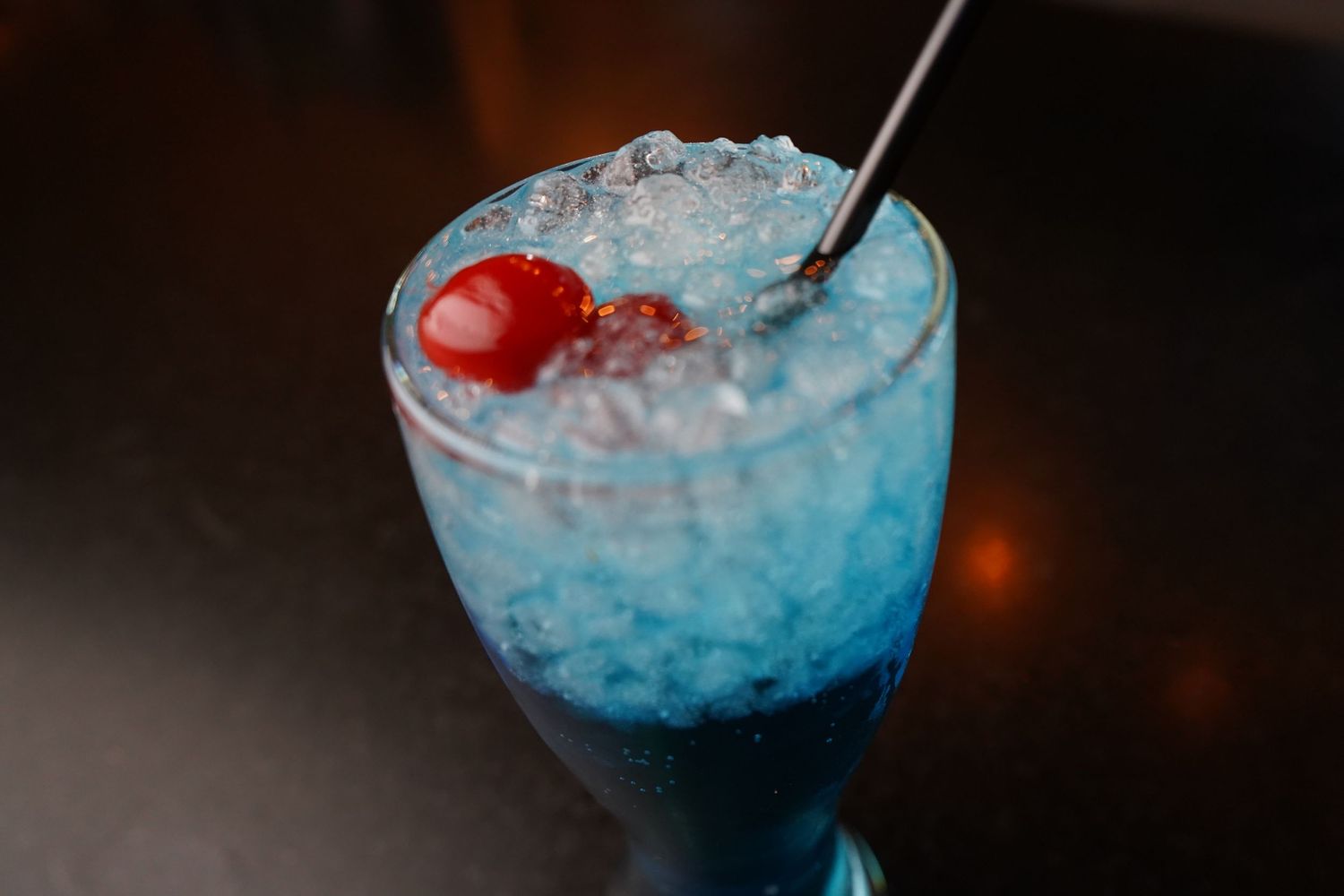 A vibrant blue cocktail with crushed ice, red maraschino cherries, and a metal straw served in a stemmed glass.