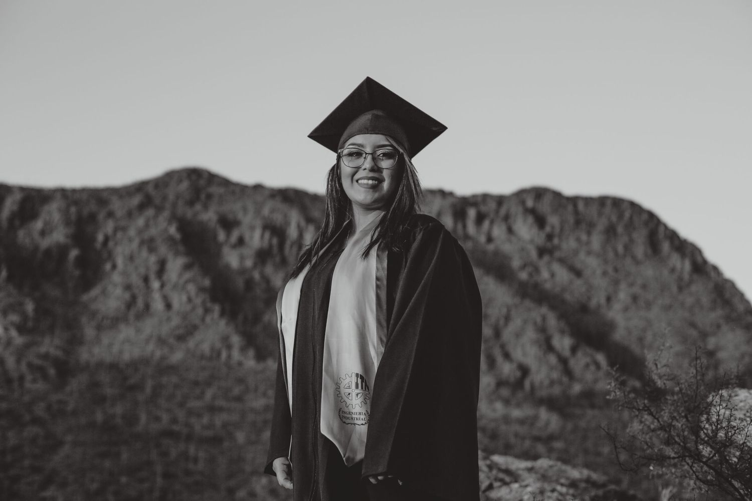 Smiling graduate in cap and gown poses outdoors with mountain backdrop in black and white photo.