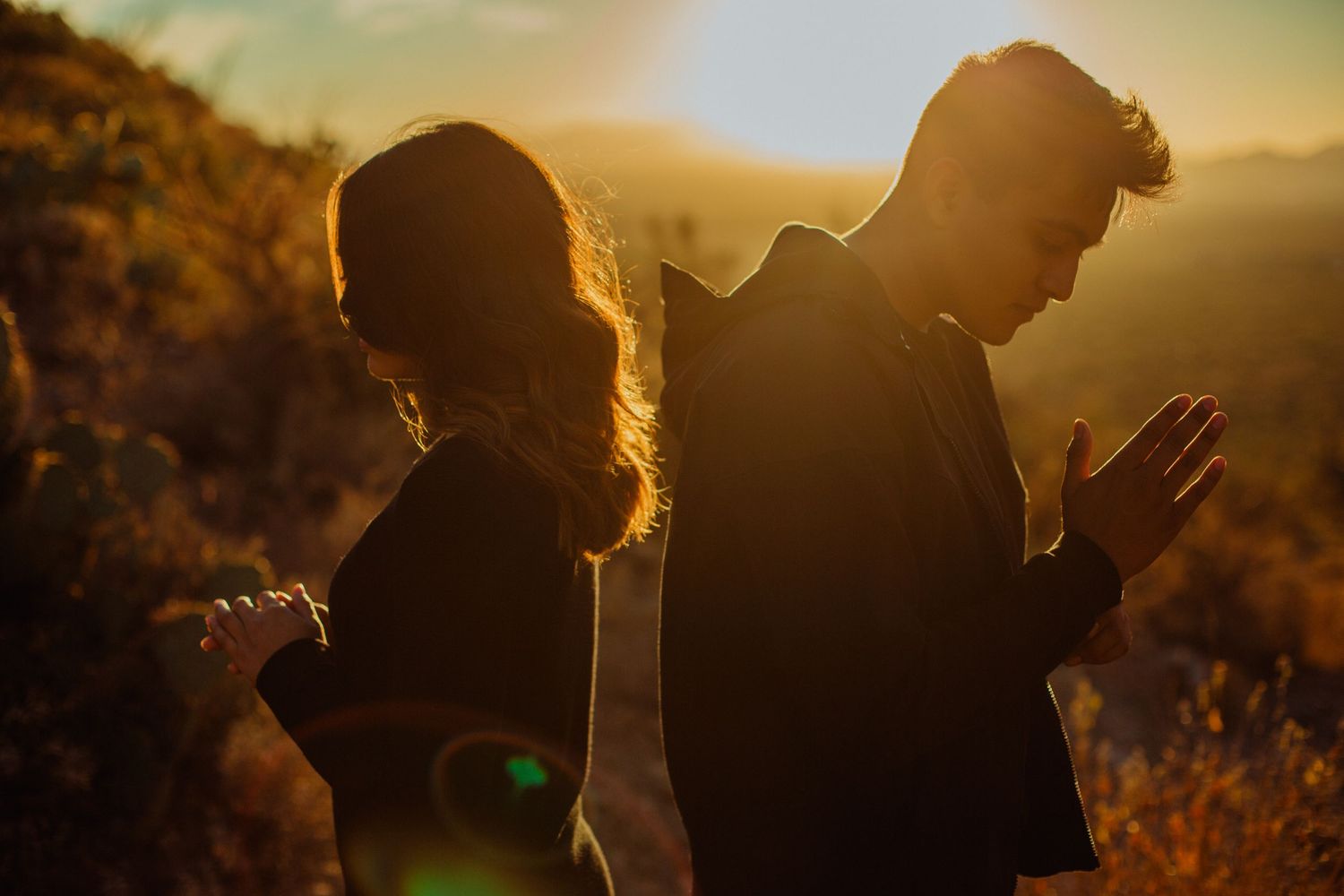 Couple standing back to back at golden sunset in mountains, silhouetted with warm glowing light behind them.