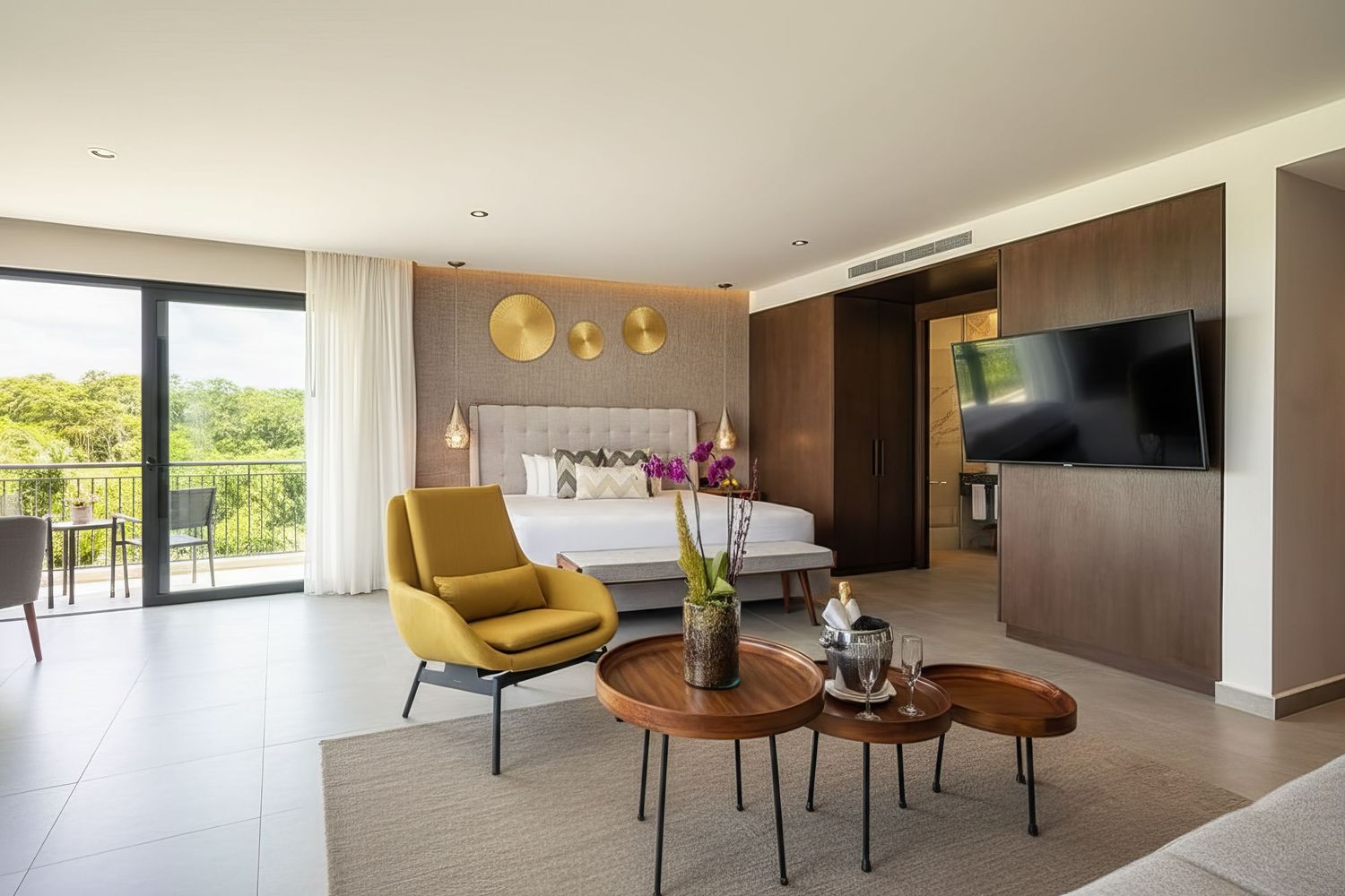 Spacious modern hotel suite featuring yellow accent chair, wooden coffee tables, flat-screen TV, and private balcony with garden views.