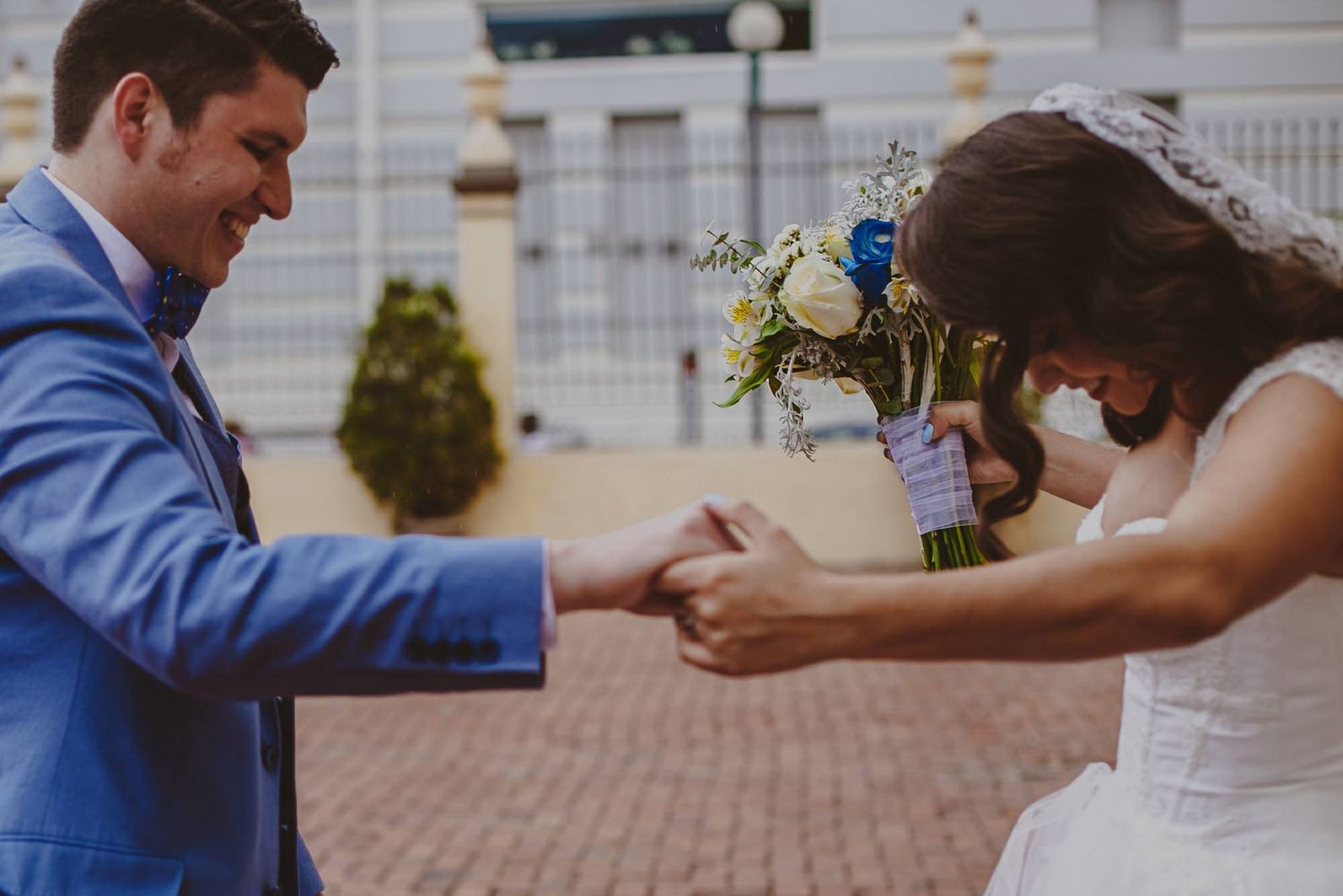Bride and groom holding hands outdoors, bride holding a white and blue floral bouquet, groom in a blue suit smiling.