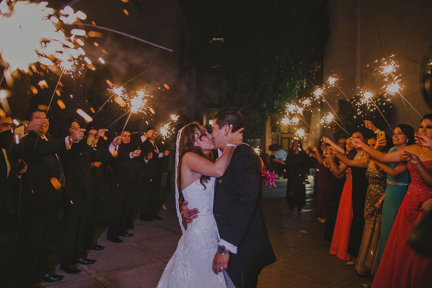 Bride and groom share a romantic kiss surrounded by guests holding sparklers at an elegant outdoor wedding celebration.