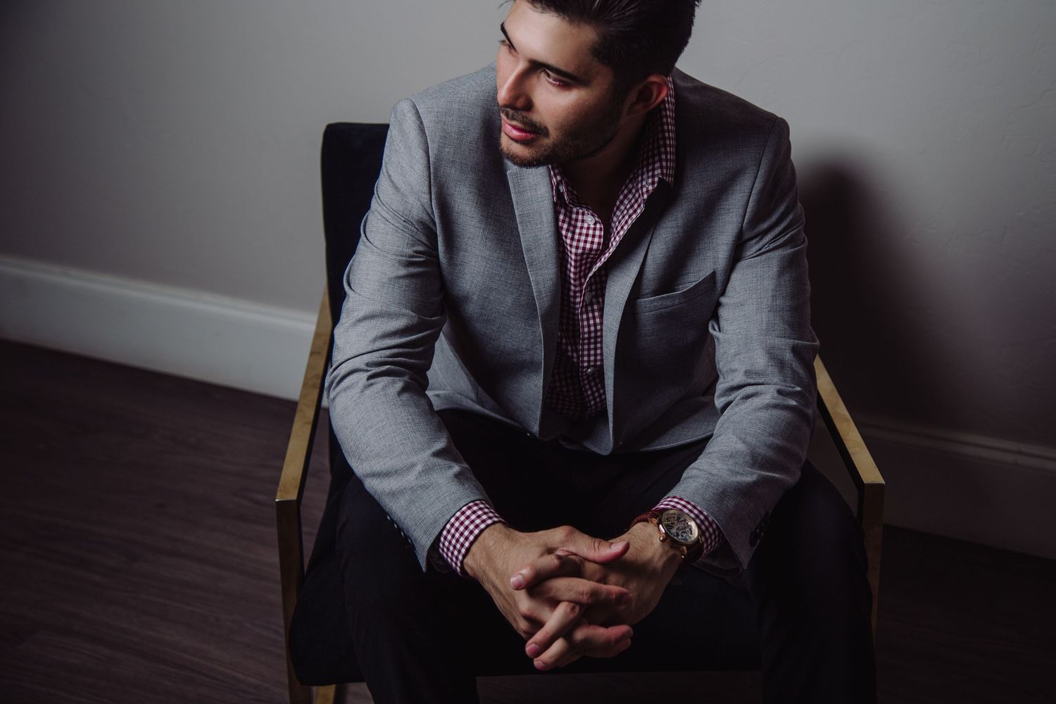 Man in gray suit sitting in chair with hands clasped, looking away thoughtfully.