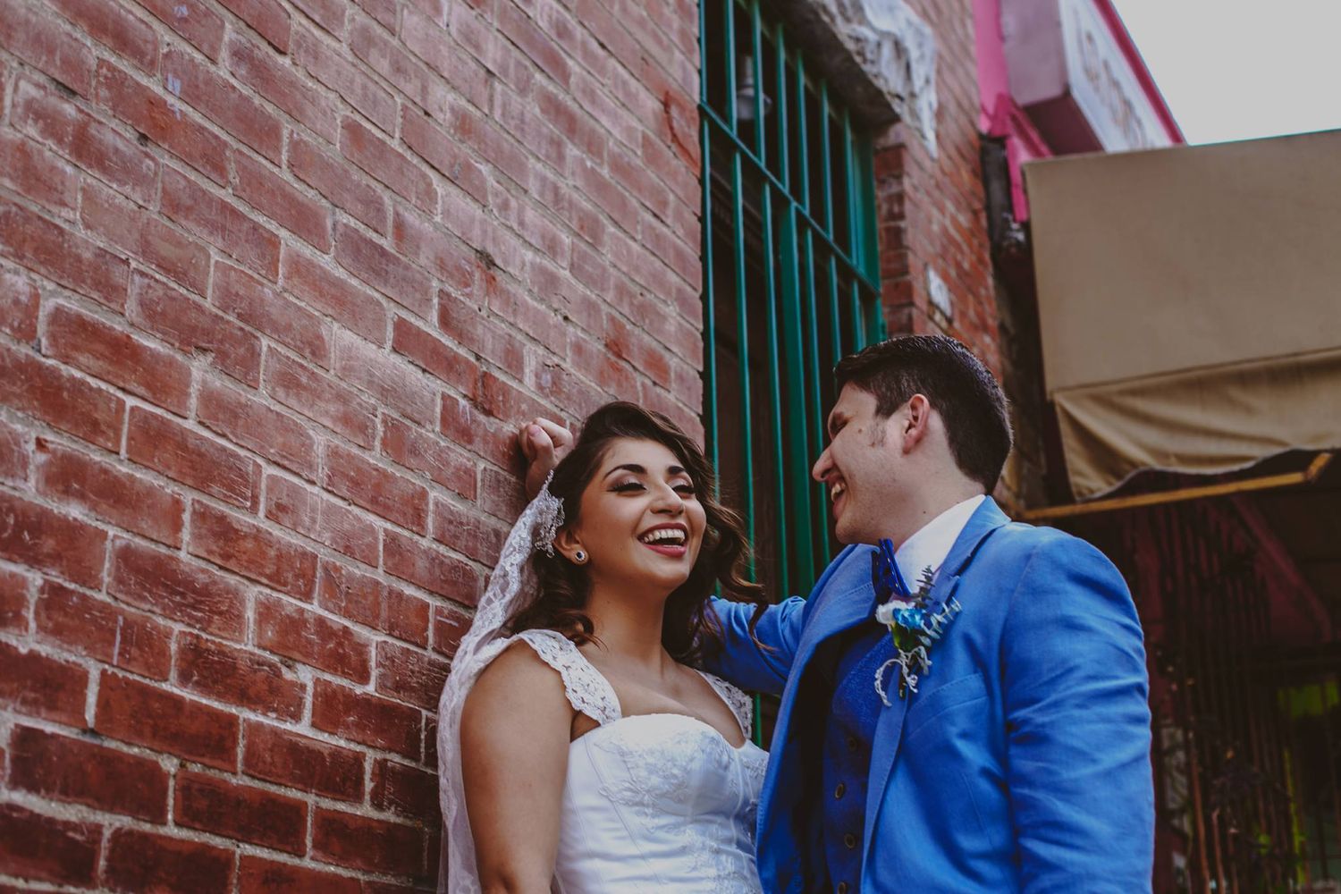 A smiling bride in a white gown and groom in a blue suit stand together against a red brick wall.