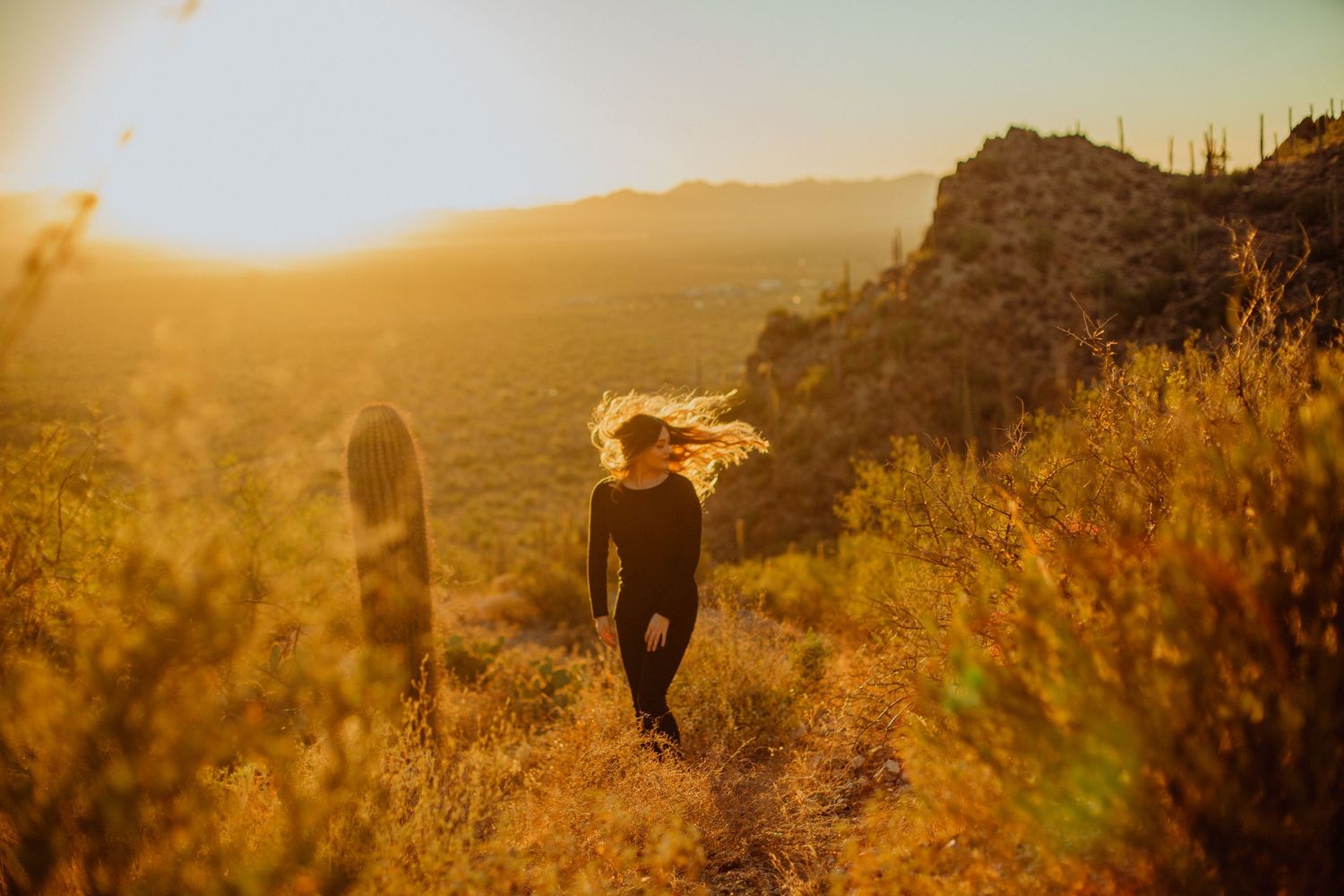 Woman in black outfit walking through golden desert landscape at sunset with wind-blown hair and cacti.