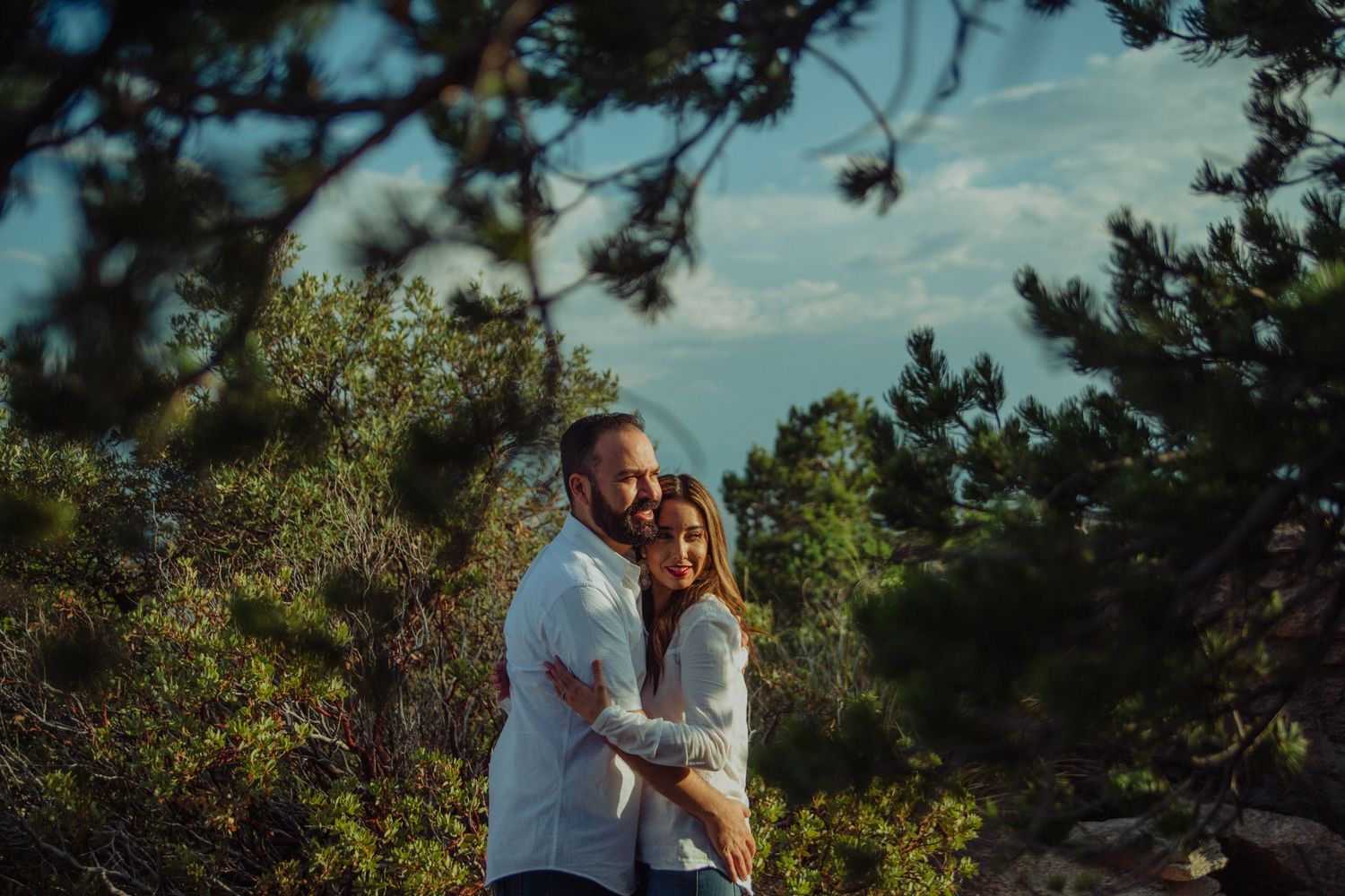 Couple embracing outdoors surrounded by lush green trees and shrubs under a blue sky.