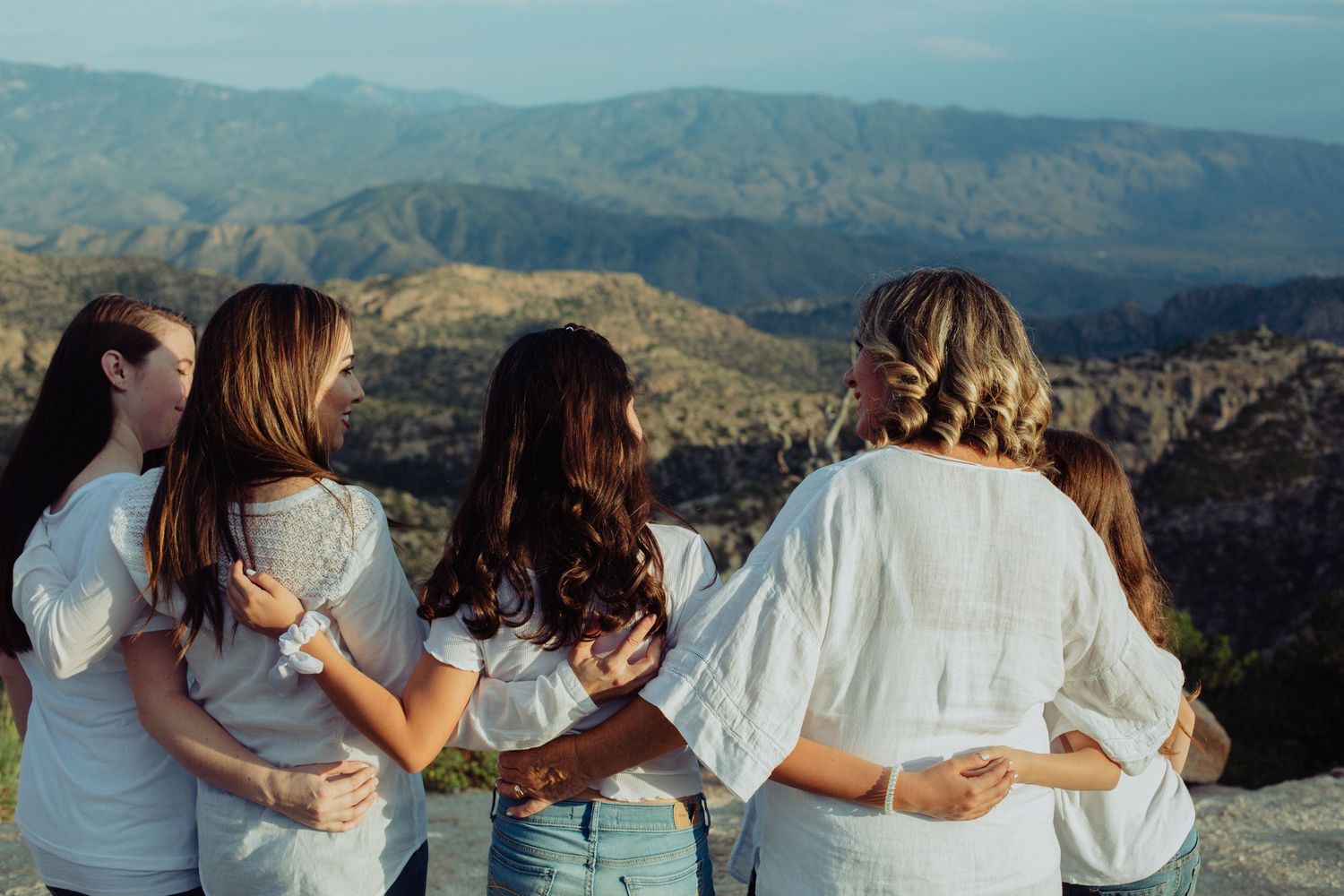 Five women in white tops stand arm in arm overlooking a scenic mountain valley landscape.