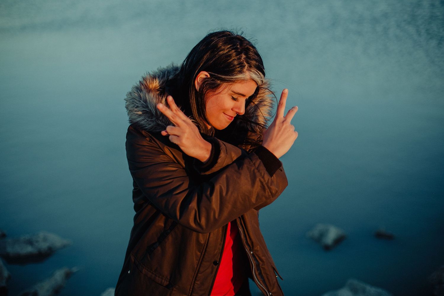 Woman in brown fur-trimmed parka making peace signs outdoors near water in golden hour light.