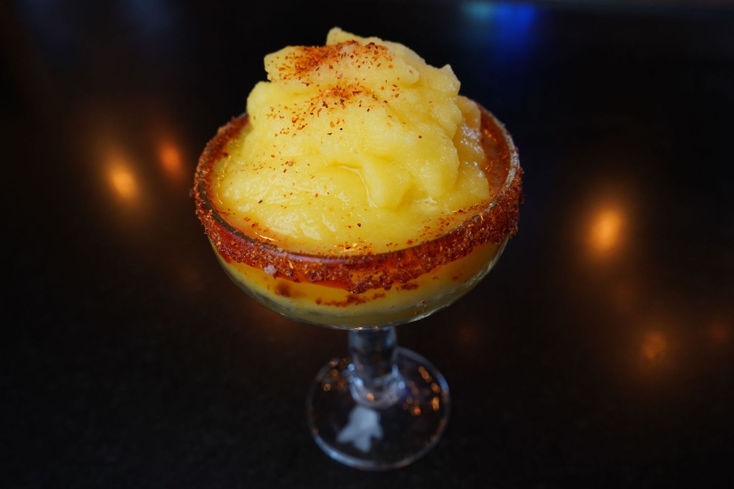 Frozen mango margarita in a chili-rimmed glass topped with shaved ice and paprika on dark background.