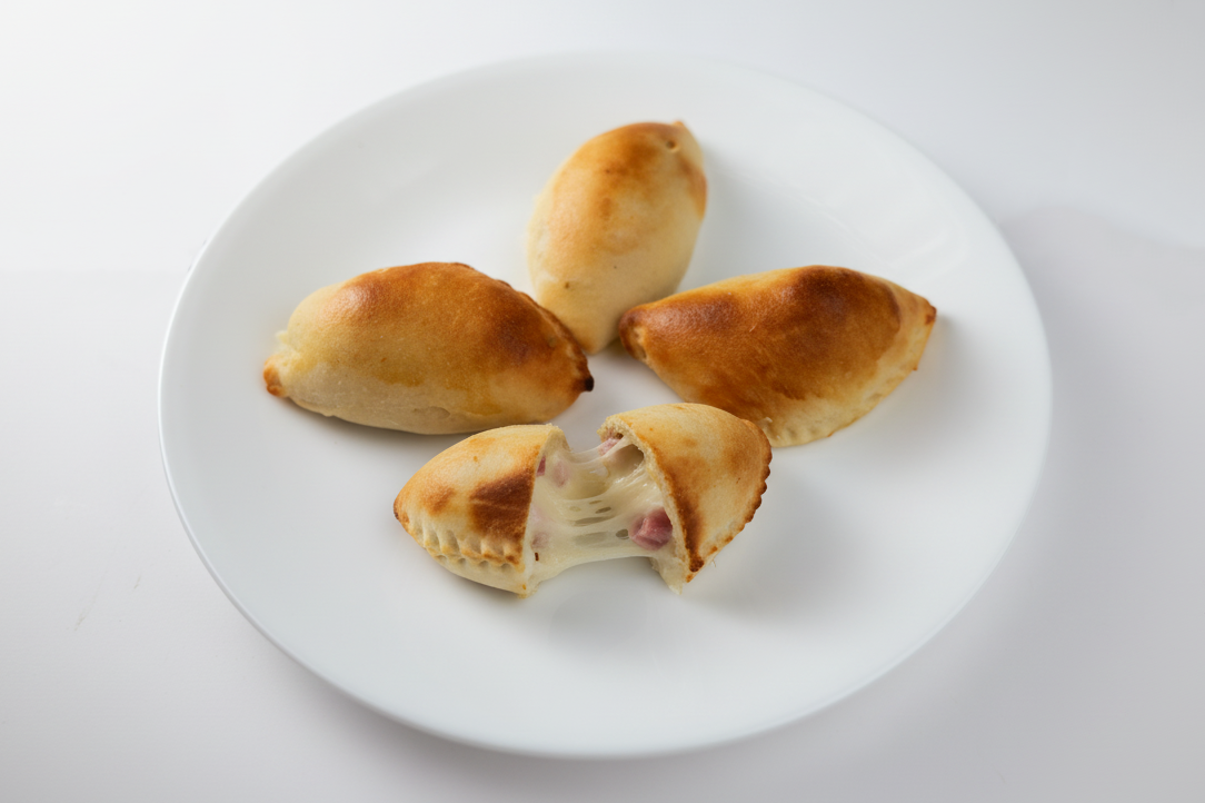 Golden-baked empanadas on a white plate, one split open revealing a savory ham and cheese filling.
