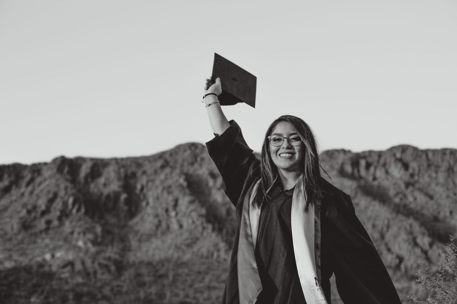 A joyful graduate in cap and gown raises her diploma outdoors with mountains in the background.