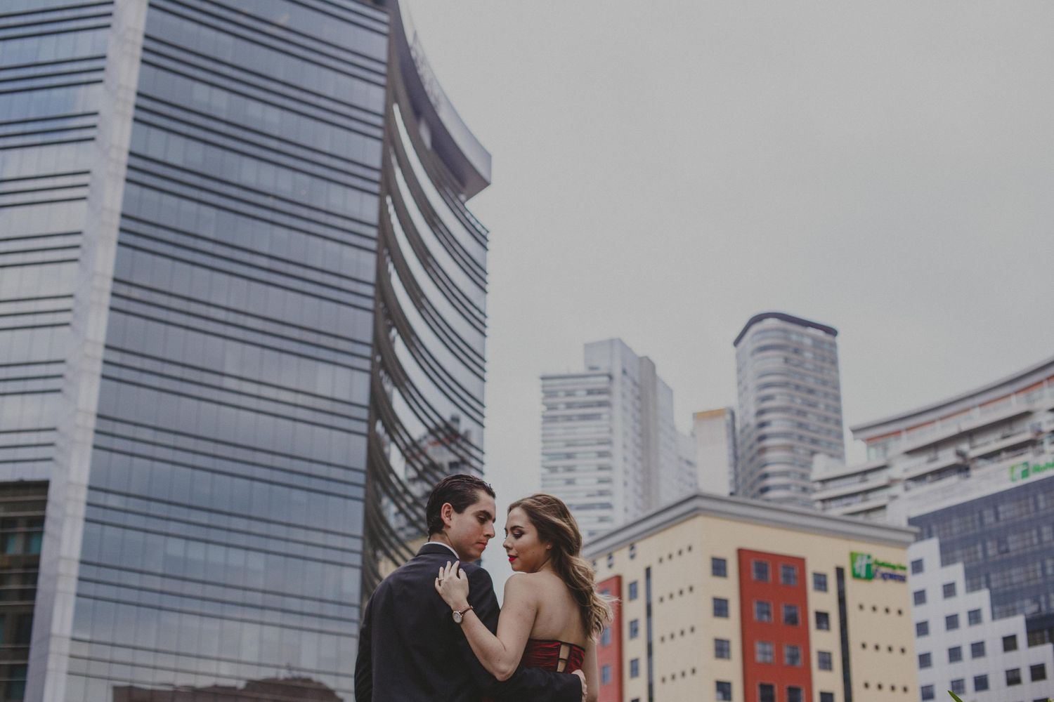Couple in formal attire sharing a romantic moment on a rooftop with modern city skyscrapers in the background.